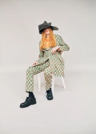 Model Filip Bryndza seated on a white chair wearing a green patterned suit and oversized dark hat against a studio backdrop.