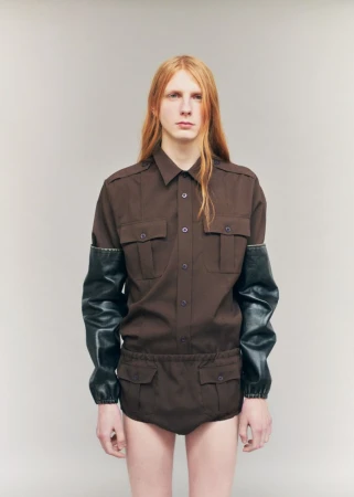 Model Filip Bryndza wearing a brown utility romper with black leather sleeves against a neutral grey studio background.