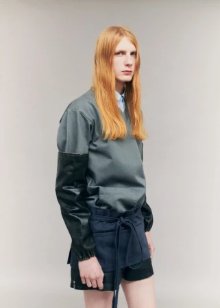 Model Filip Bryndza with long red hair wearing a grey top, black leather sleeves, and navy wrap against a light grey studio background.