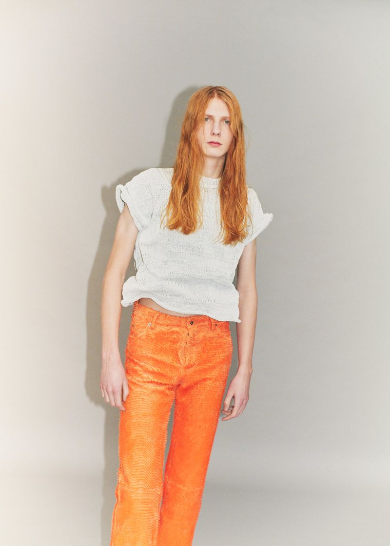 Model Filip Bryndza with long red hair wearing a white textured top and bright orange trousers against a grey studio backdrop.