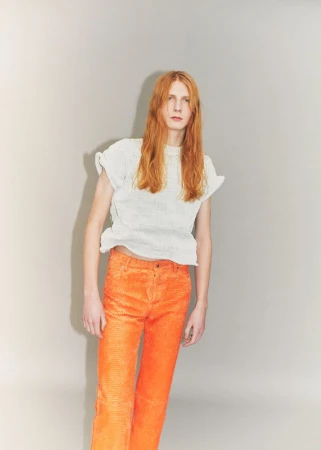 Model Filip Bryndza with long red hair wearing a white textured top and bright orange trousers against a grey studio backdrop.