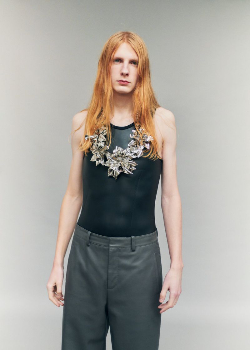 Model Filip Bryndza with long red hair wearing a black tank top, slate trousers, and a sculptural silver necklace against a grey backdrop.
