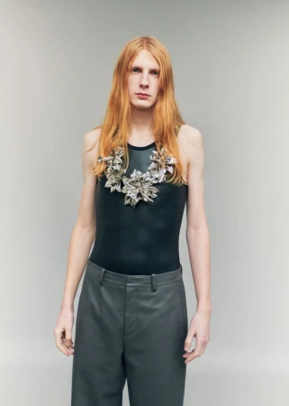 Model Filip Bryndza with long red hair wearing a black tank top, slate trousers, and a sculptural silver necklace against a grey backdrop.