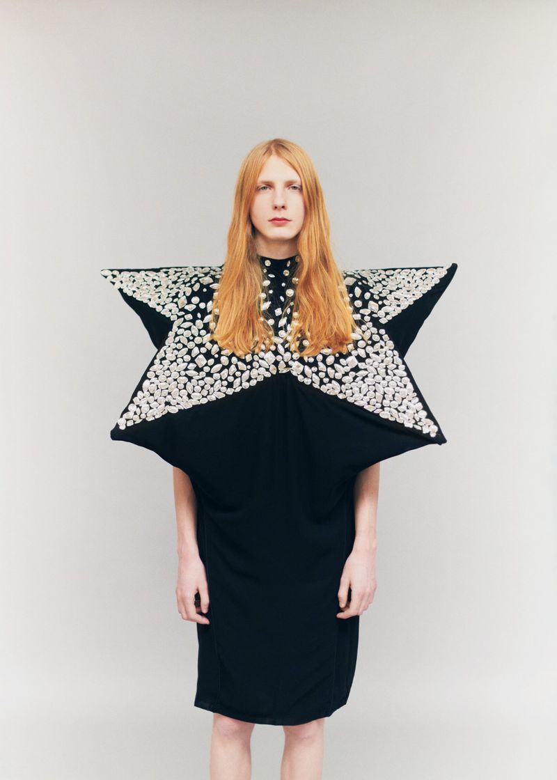 Model with long reddish-blonde hair wearing a sculptural star-shaped black dress with silver embellishments against a plain grey backdrop.
