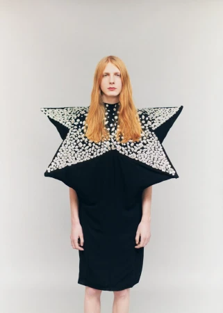 Model with long reddish-blonde hair wearing a sculptural star-shaped black dress with silver embellishments against a plain grey backdrop.