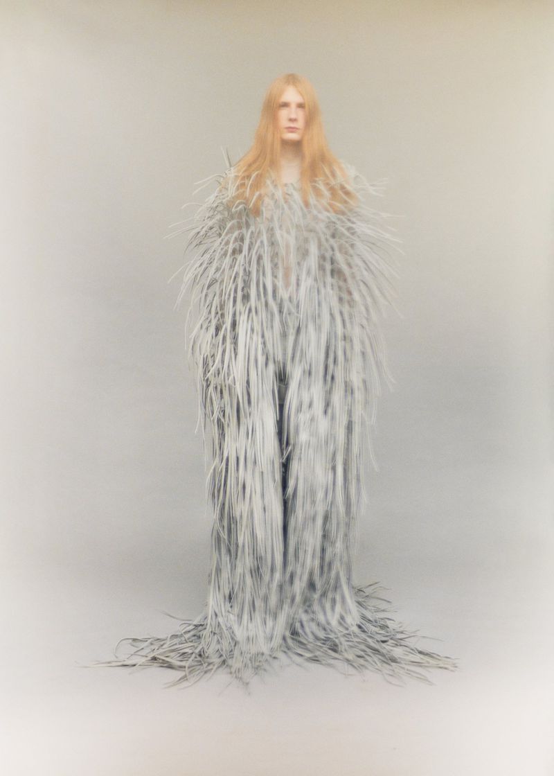 Model with long blonde hair wearing a floor-length feathered grey garment against a plain neutral backdrop