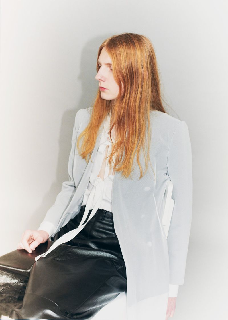 Model Filip Bryndza with long red hair wearing a light grey blazer, tied white blouse, and black leather trousers seated against a pale wall.
