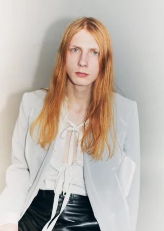 Filip Bryndza with long red hair wearing a pale blue blazer, white ribbon-tied blouse, and black leather trousers.