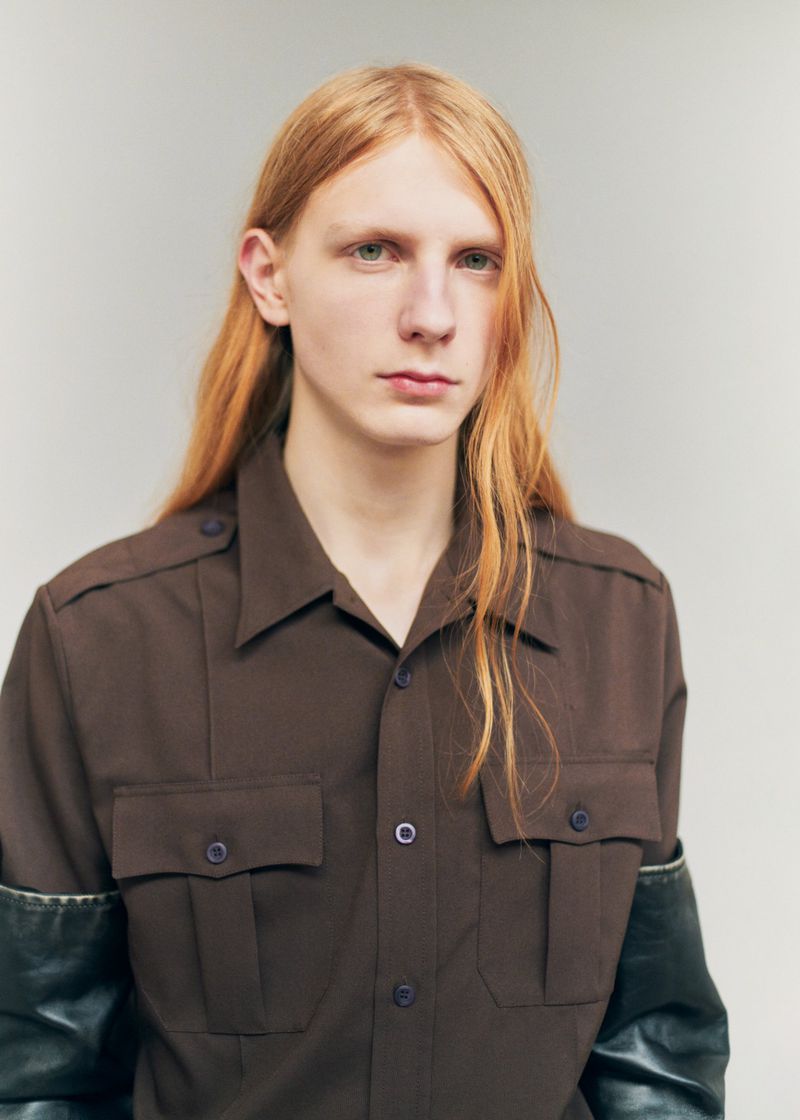 Portrait of a male model with long red hair wearing a dark brown button-down shirt against a neutral background
