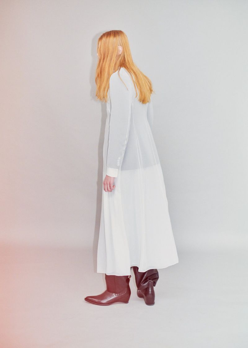 Model Filip Bryndza seen from behind in a long white layered dress and brown leather boots against a neutral grey studio backdrop.