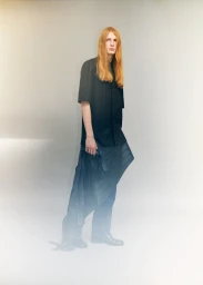 Model Filip Bryndza with long strawberry blonde hair wearing an all-black shirt and pleated wrap trousers in a minimal studio setting.