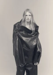 Model Filip Bryndza wearing an oversized draped black leather garment against a plain grey studio backdrop.