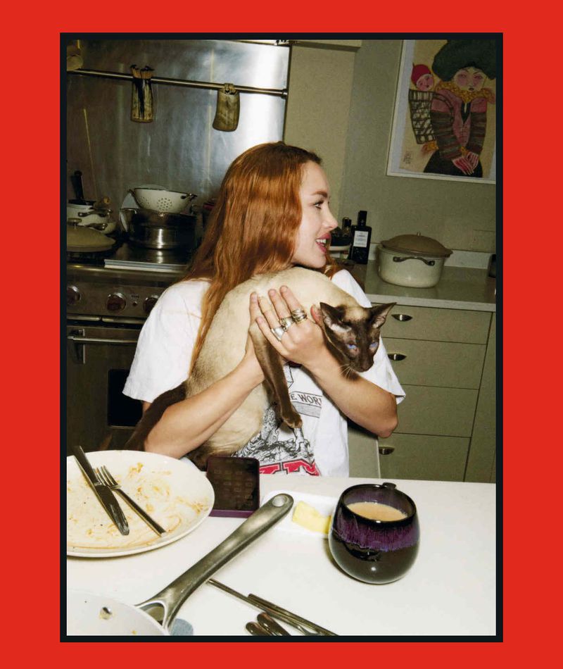 Red-haired woman seated at a kitchen table holding a Siamese cat near a plate and mug.