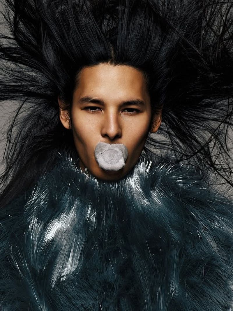 Portrait of a model with windblown black hair holding a pacifier in his mouth and wearing a voluminous dark teal fringed collar.