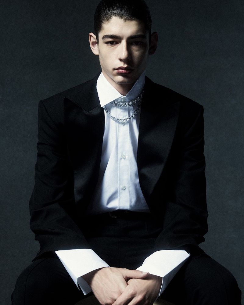 Model Baptiste Vivo wearing a black tuxedo jacket, crisp white shirt, and crystal chain necklace against a dark background.
