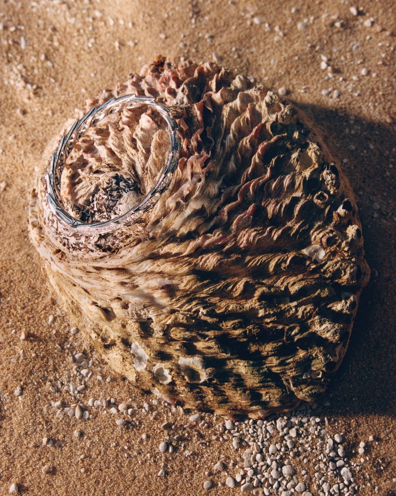 Silver bangle bracelet resting on a large textured seashell surrounded by coarse sand and small pebbles in warm directional light.