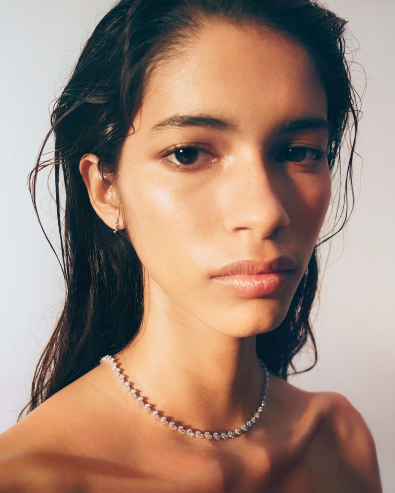 Close-up portrait of a woman with wet hair wearing a diamond necklace and small earring against a pale background.