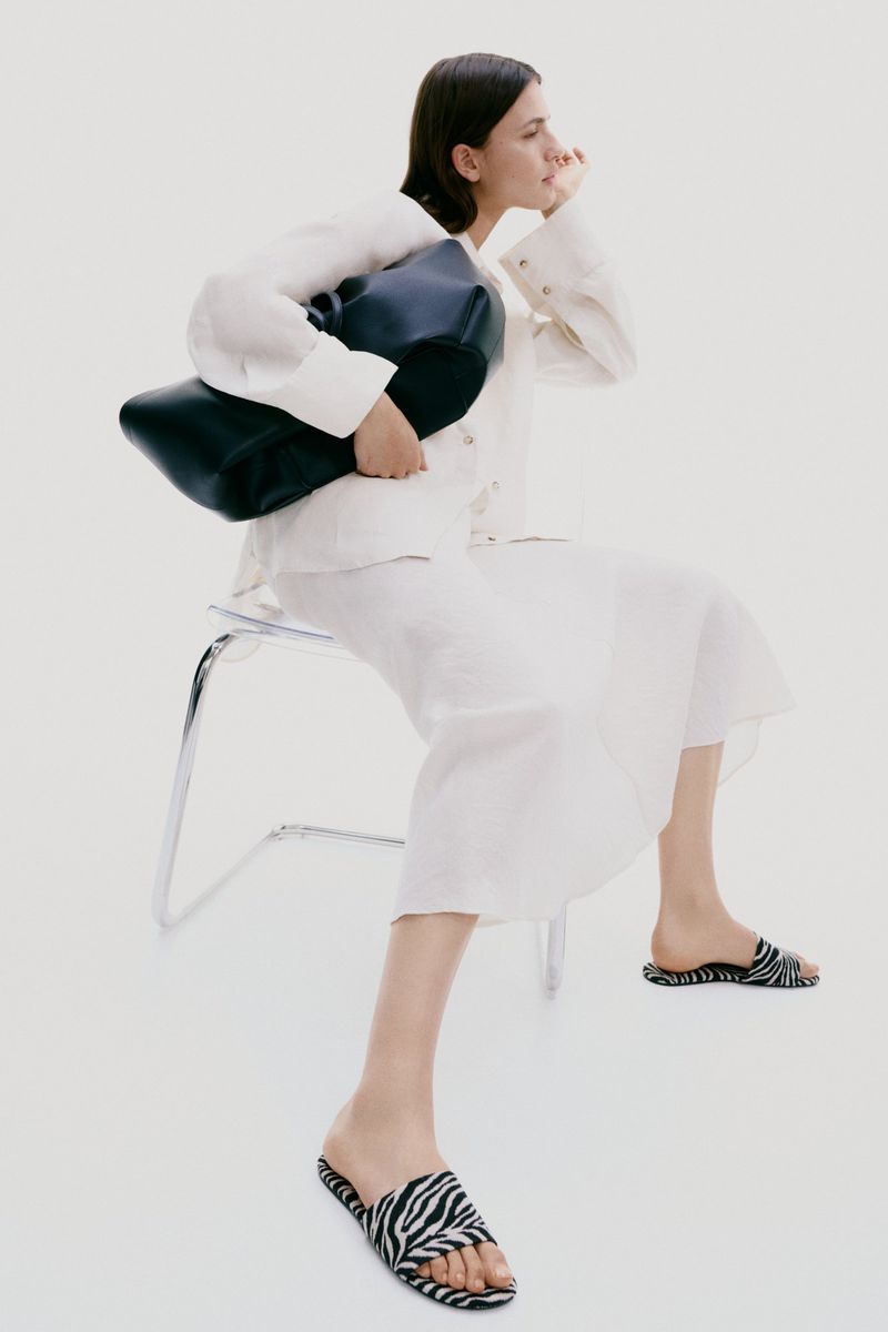 A woman in a white linen skirt and top sits on a transparent chair holding a black purse and wearing zebra-striped sandals in a studio.