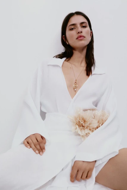 Brunette woman in an unbuttoned white shirt dress with a flower-like accessory, posing in a bright studio against a white wall.