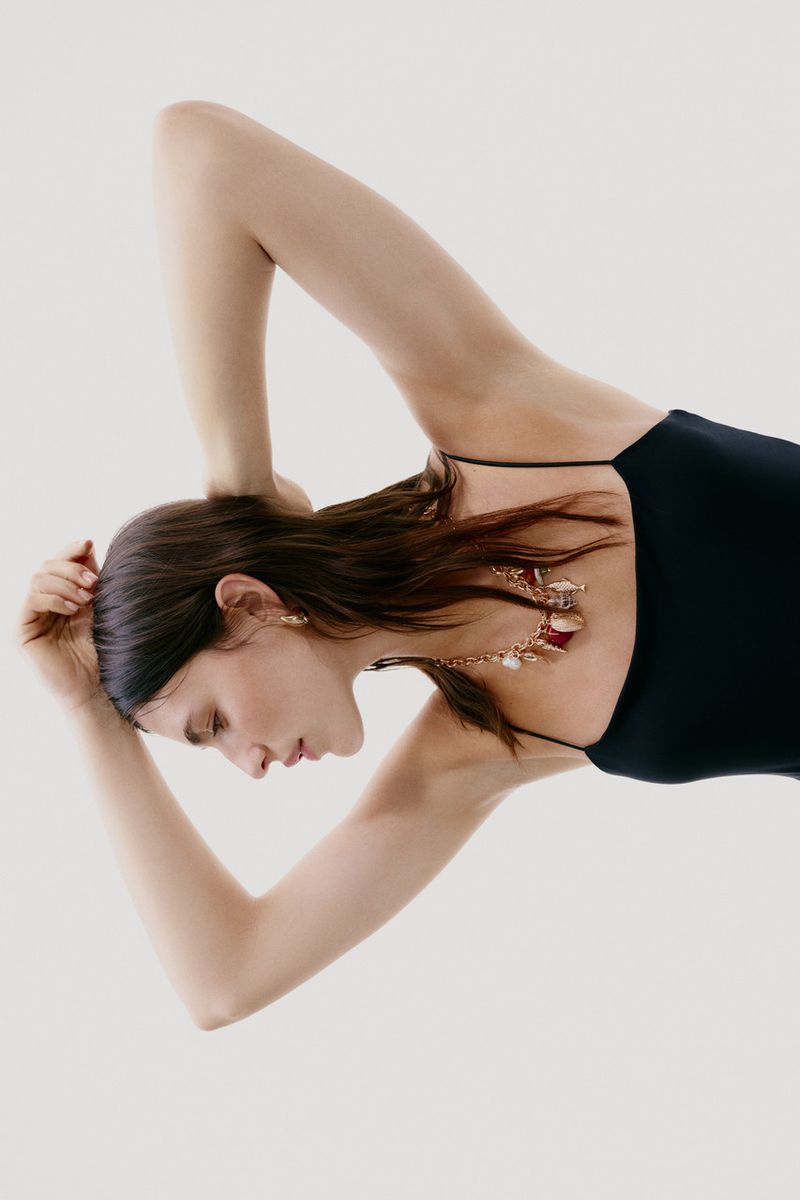 Woman with brown hair and black top lies with arms raised to show a chain necklace with red and gold pendants on a white background.