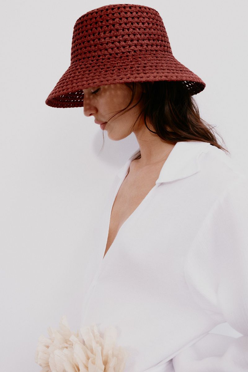 Portrait of a stylish woman wearing a red woven sun hat and a white collared shirt against a plain background in soft lighting.