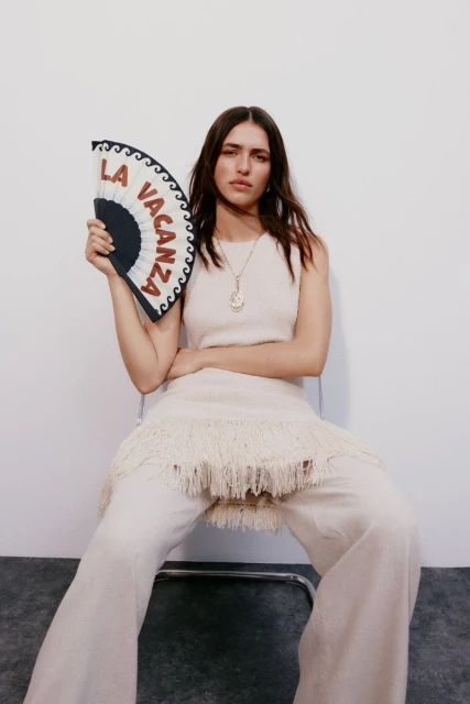 A brunette woman sits on a chair in a fashion-style full shot, wearing a cream knit outfit and holding a decorative fan against a white background.