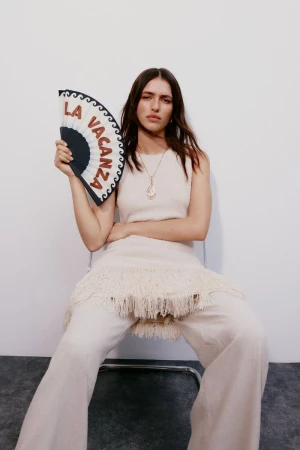 A brunette woman sits on a chair in a fashion-style full shot, wearing a cream knit outfit and holding a decorative fan against a white background.