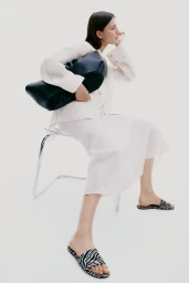 Model seated on a clear chair wearing a white shirt and skirt, holding a large dark handbag against a white background