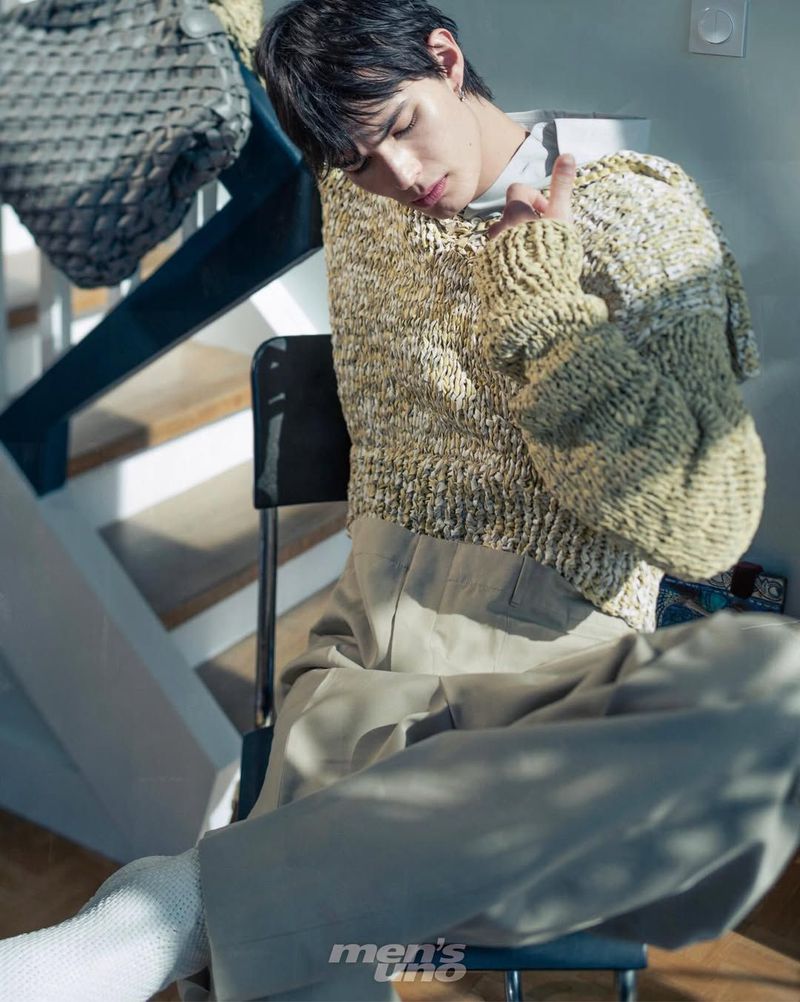 A young man seated in a chair by an indoor staircase wearing a chunky marl knit sweater and wide beige trousers.