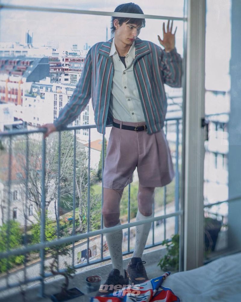 A young male model in a striped jacket and mauve pleated shorts standing on a balcony overlooking Parisian rooftops through a window.