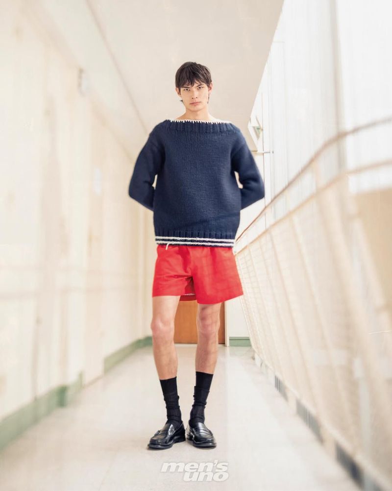 Male model posing in a bright hallway wearing an oversized navy boatneck sweater, red tailored shorts, and black leather loafers.