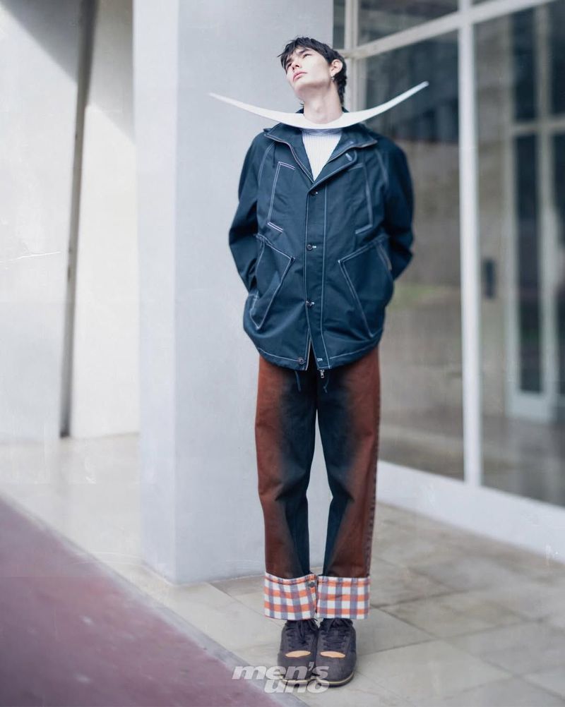 Model wearing a navy contrast-stitch jacket, ombre cuffed trousers, and a sculptural curved white collar accessory against glass windows.