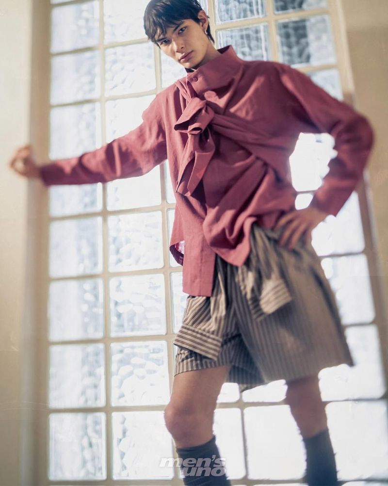 Male model wearing a draped mauve long-sleeve shirt and striped shorts posing in front of a large grid window.