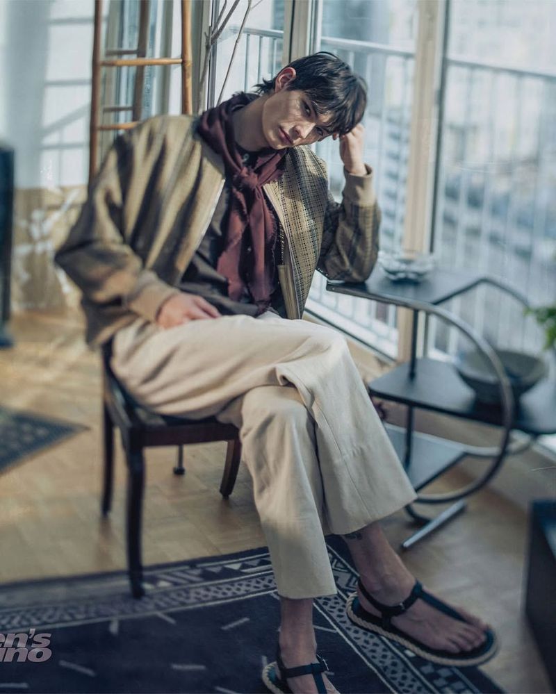 Model seated on a chair by a window wearing a beige jacket, burgundy neckwear, wide-leg cream trousers, and strap sandals.