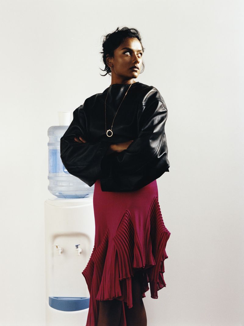 Simone Ashley in a black voluminous top and pleated magenta skirt poses beside an office water cooler against a white studio backdrop.