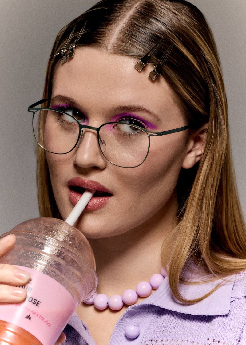 A young woman wearing wire-frame glasses, silver hair clips, and a lilac knit top sips a pink beverage through a straw.
