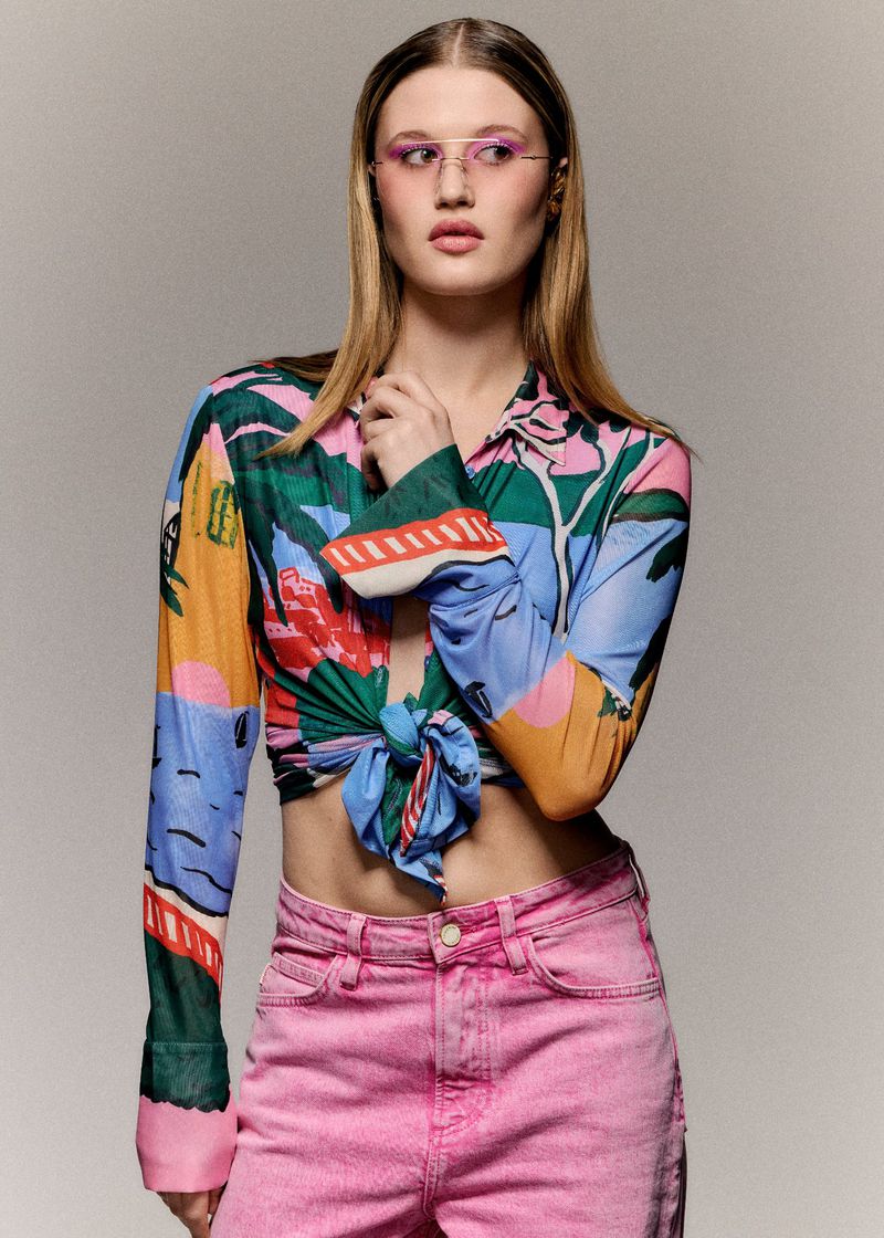 Model wearing a colorful tied graphic crop shirt and pink jeans against a neutral studio backdrop.