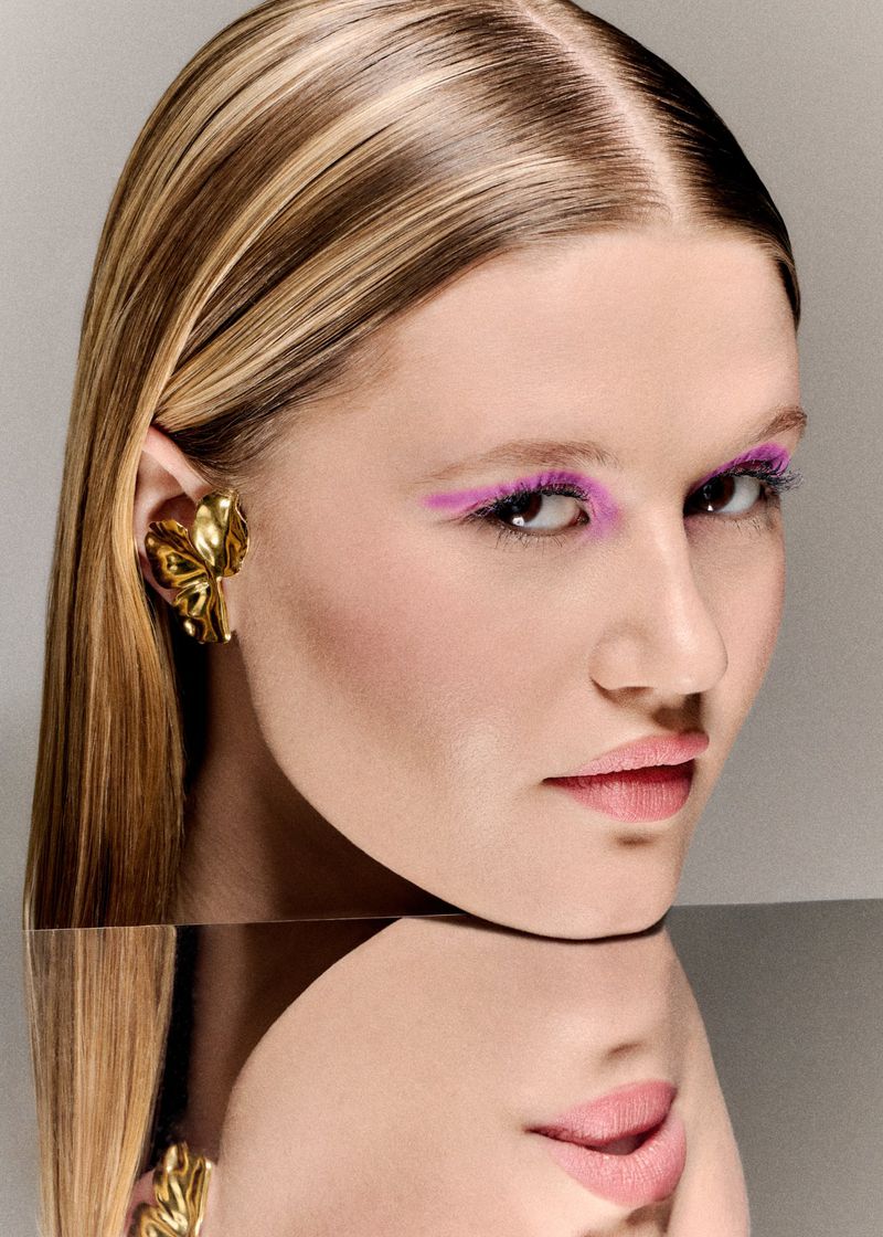 Close-up portrait of a woman with pink eyeshadow, sleek hair, and a gold butterfly ear cuff reflected in a mirror below.