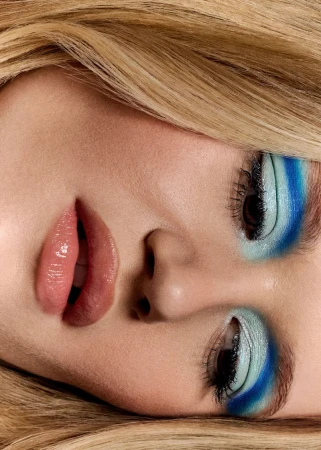 Extreme close-up beauty portrait of a woman with blonde hair, glossy pink lips, and vibrant layered blue eye makeup.
