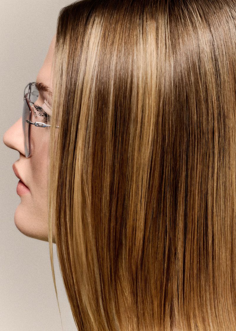 Profile view of a woman with sleek straight highlighted hair wearing tinted rimless sunglasses against a neutral studio backdrop.