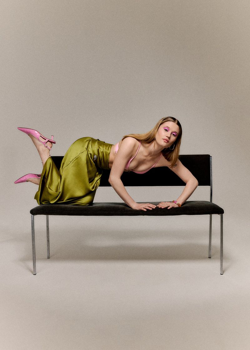 Model wearing an olive green satin skirt and pink slingback heels poses across a black bench in a studio setting.