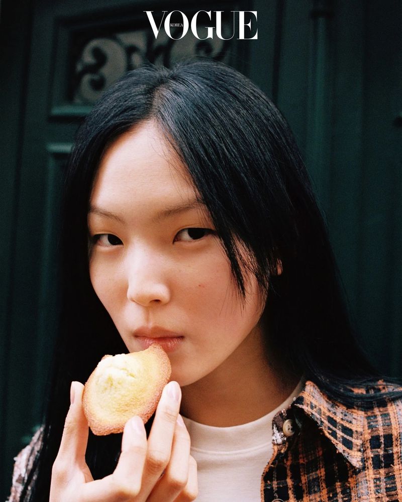 Close-up portrait of model Yun Seo Woo holding a baked madeleine pastry to her lips in front of a dark green paneled door.