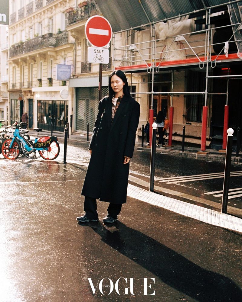 Model Yun Seo Woo wearing a long black overcoat standing on a sunlit wet street in Paris.
