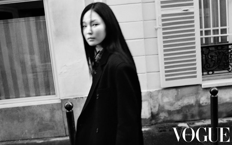 Model Yun Seo Woo wearing a dark tailored coat walking along an urban street with window shutters in black and white.