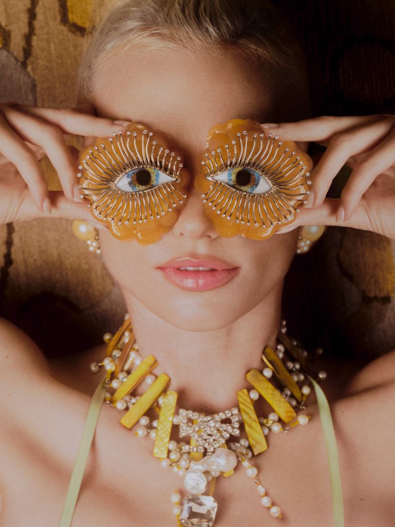 Close-up of a blonde woman holding ornate eye-shaped ornaments over her eyes, wearing an elaborate yellow statement necklace.