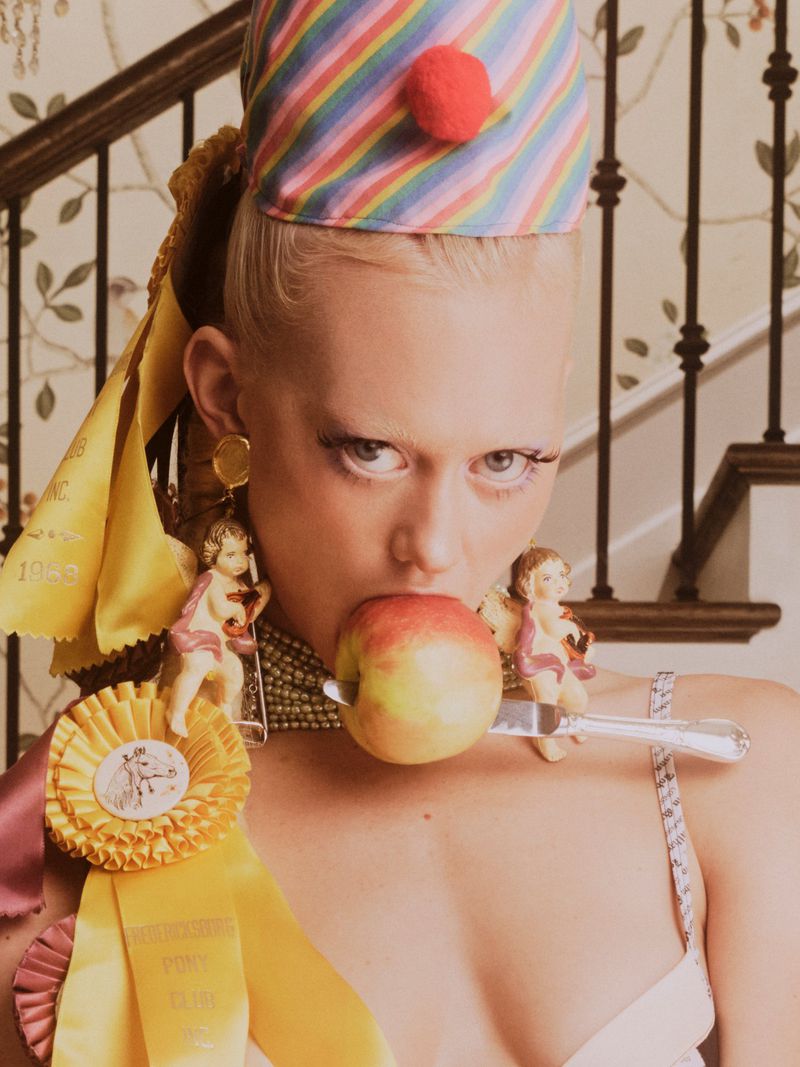 Close-up portrait of a woman wearing a rainbow party hat, angel earrings, and holding an apple pierced by a knife in her mouth.