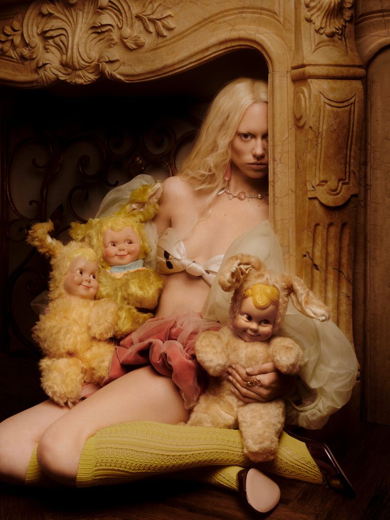 Blonde woman sitting inside an ornate carved fireplace holding vintage plush toys with doll faces for Ladygunn Magazine editorial.