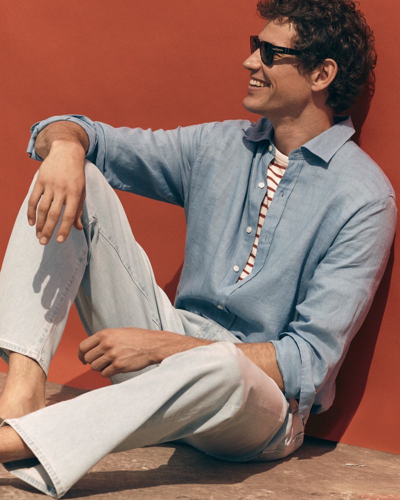 Model Paul Hupfer seated against a terracotta wall wearing sunglasses, a light blue linen shirt, and light-wash denim jeans.