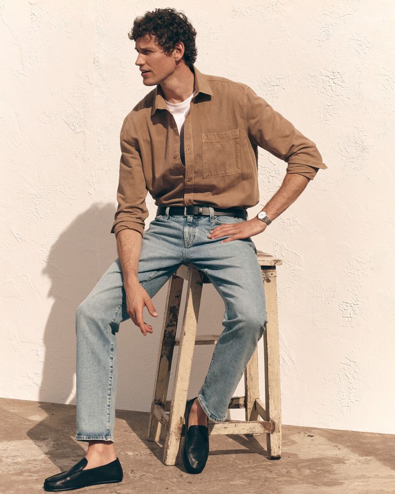 Model Paul Hupfer seated on a wooden stool wearing a brown button-up shirt and light wash blue jeans against a sunlit wall.