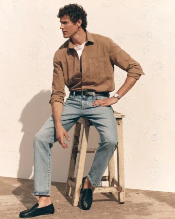 Model Paul Hupfer seated on a wooden stool wearing a brown button-up shirt and light wash blue jeans against a sunlit wall.